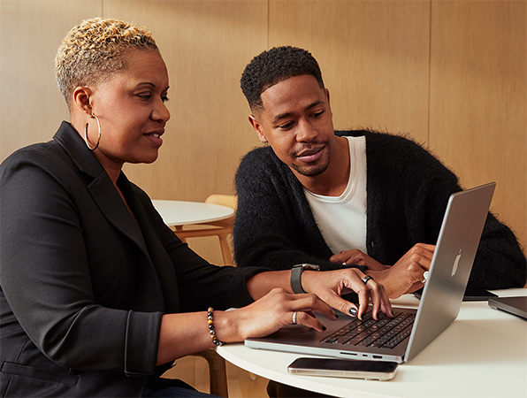 Dwie osoby z personelu Apple siedzą przy stole i pracują wspólnie na MacBooku. 