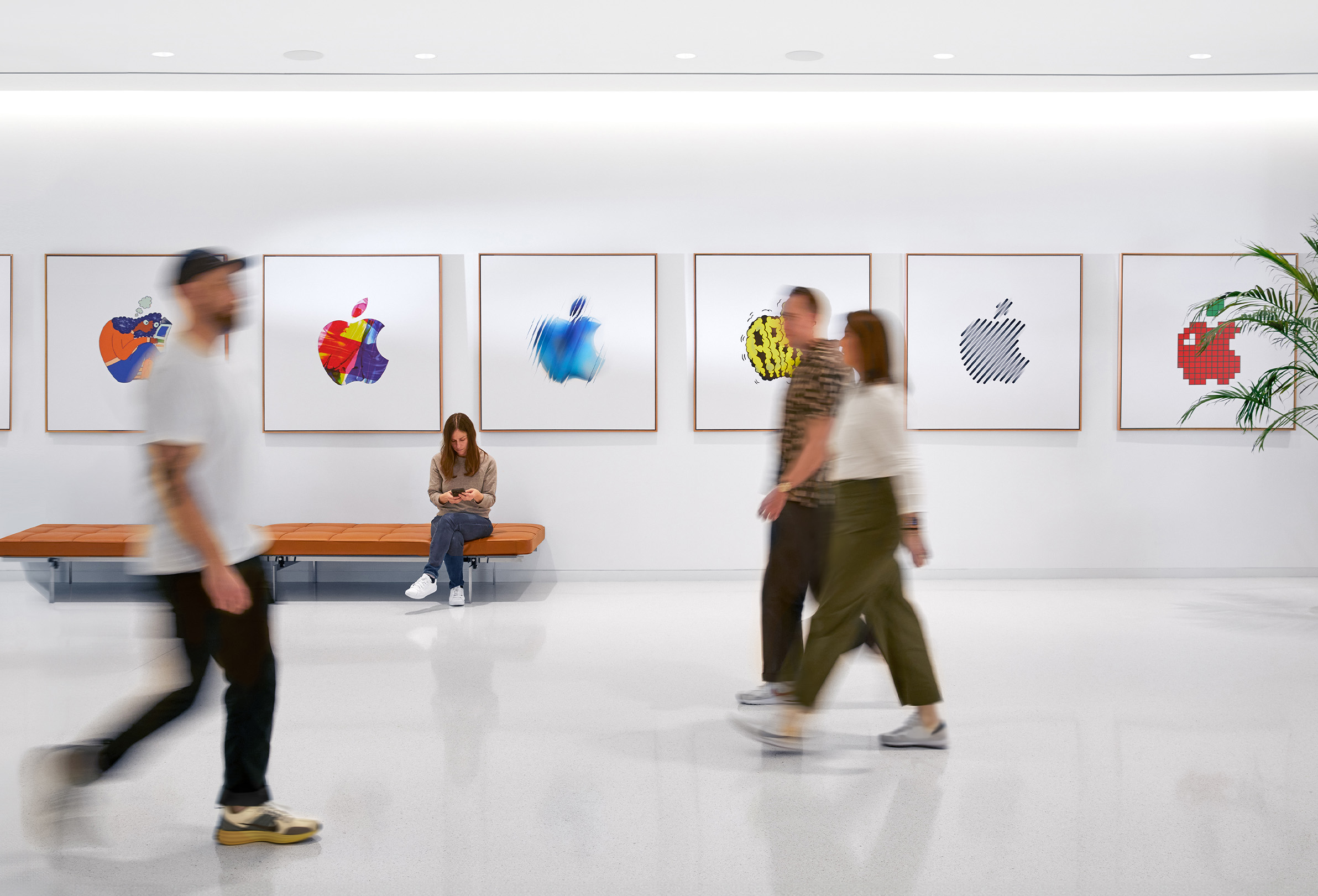 Trzy osoby z personelu Apple idą korytarzem, na ścianach widać galerię kolorowych logo Apple. Czwarta osoba siedzi na sofie.