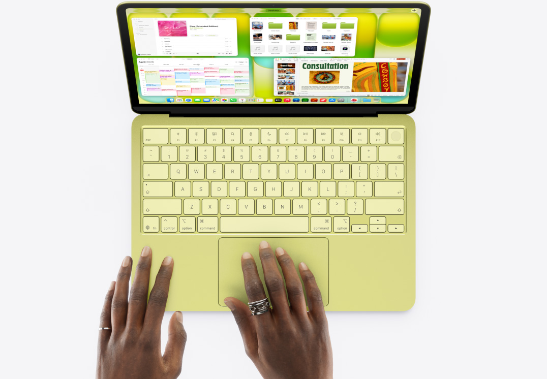 MacBook Neo, warna citrus, tangan seseorang di trackpad, beberapa jendela terbuka termasuk aplikasi Musik, Kalender, dan Microsoft PowerPoint
