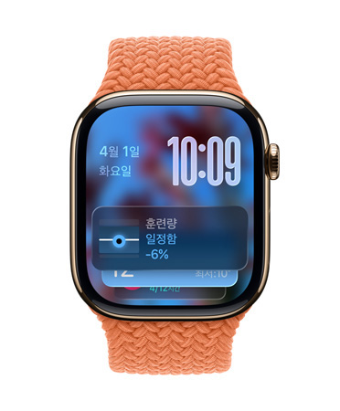 탠저린 브레이드 밴드가 장착된 로즈 골드 Apple Watch, 왼쪽 상단의 날짜가 표시된 시계 페이스, 오른쪽 상단의 겹쳐진 상태로 표시된 시간, 하단의 훈련량 위젯, 모두 반투명 Liquid Glass 디자인이 적용된 모습