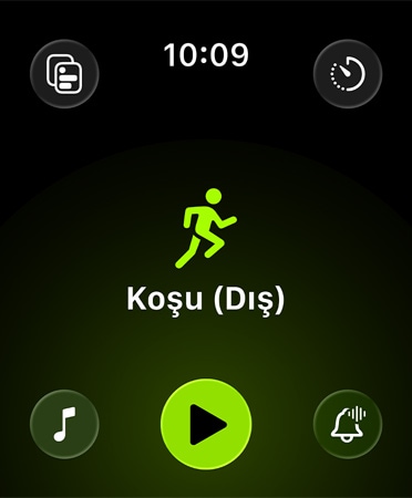 Antrenman uygulamasının yeni görünümü. Siyah arka plan üzerinde ekranın her köşesine yerleştirilmiş yeşil düğmeler; uyarılar, müzik, deneyimler, özel antrenmanlar ve ölçümlere erişim sağlıyor. Ortada yeşil renkli bir koşucu simgesi, altında beyaz harflerle Koşu (Dış) yazısı, en altta yeşil bir Oynat düğmesi yer alıyor