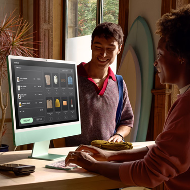 En medarbejder sidder bag disken i en surfbutik og bruger en iMac til at gennemføre et køb for en kunde på den anden side af disken.