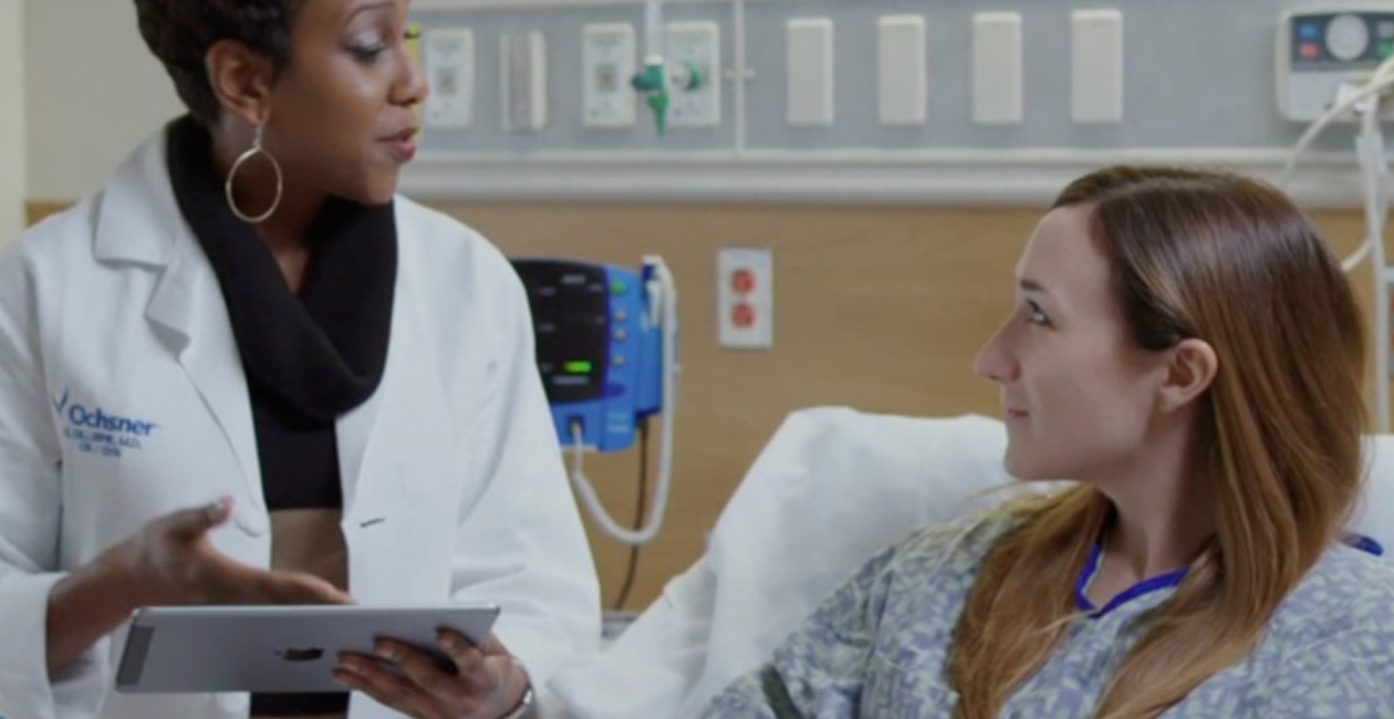 Uma profissional de saúde com um iPad falando com uma paciente em uma cama de hospital.