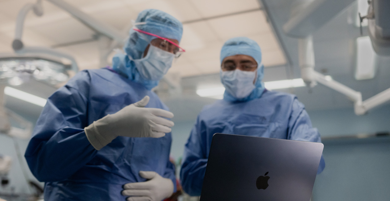 Dois cirurgiões falando sobre algo na tela de um MacBook Pro na sala de cirurgia.