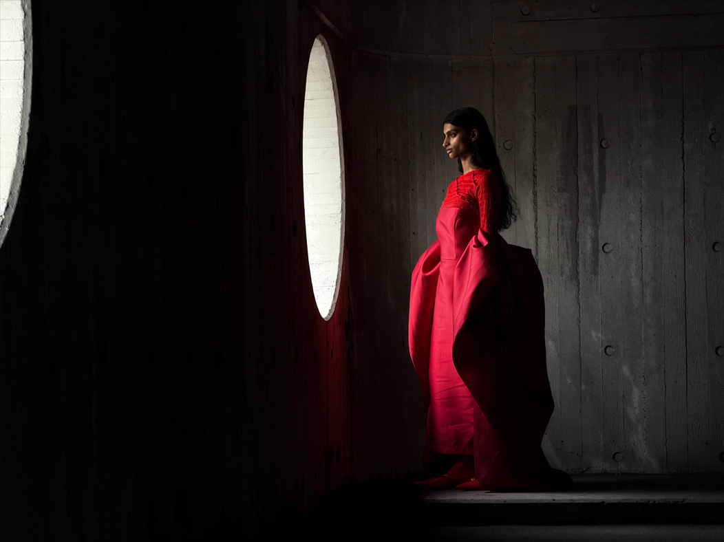 Uma mulher com um longo vestido vermelho a ser fotografada numa sala vazia. A app Fotografia muda de distância focal para ilustrar as capacidades do sistema de câmaras Pro, a mostrar a opção de 100 mm