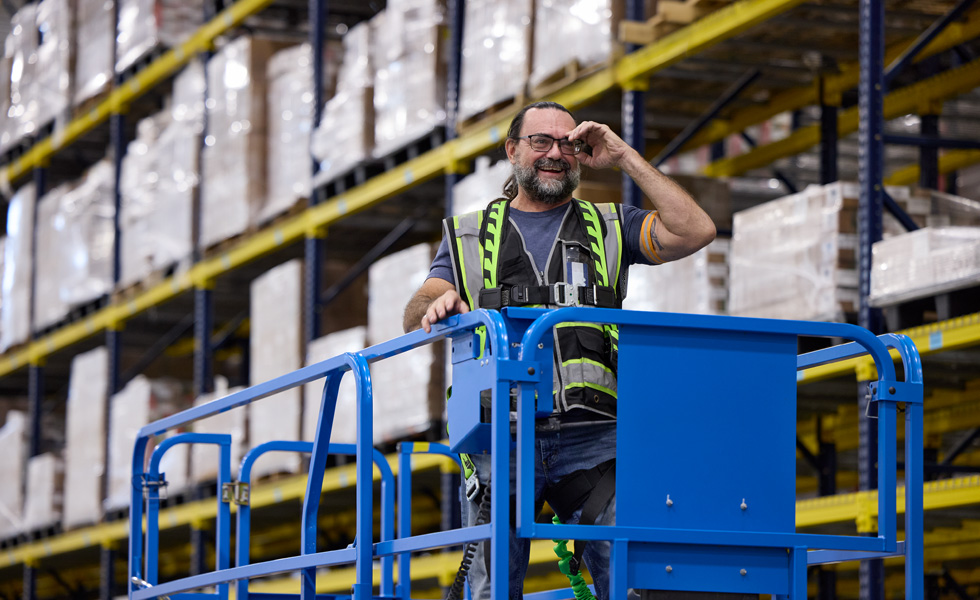 A warehouse employee wearing a high-visibility vest rides a driveable lift