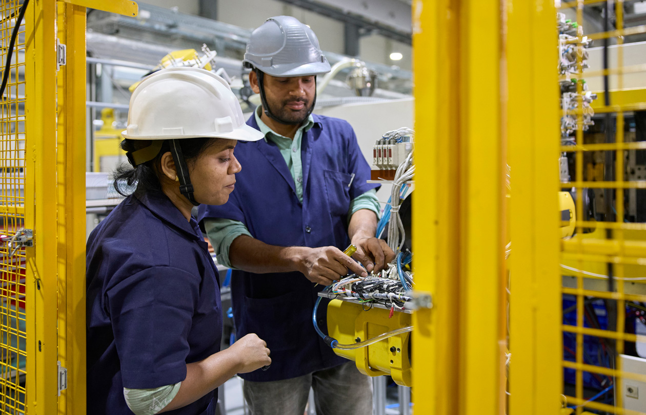 Two employees in work smocks and hats stand at their workstations operating assembly machines
