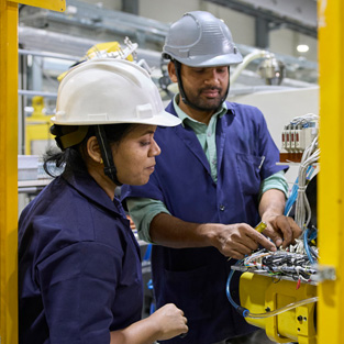 Two employees in work clothes and safety hats stand in front of an assembly machine