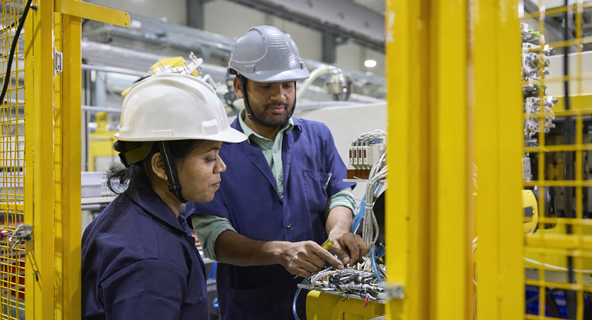 Two employees in work clothes and safety hats stand in front of an assembly machine